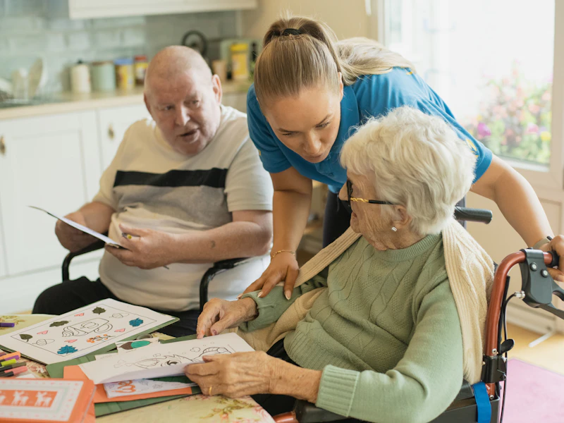 Une aide à domicile accompagne une dame âgée en fauteuil roulant et un monsieur âgé, autour d'une table.