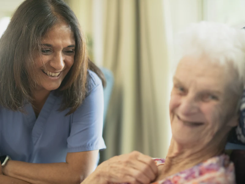 Une soignante souriante, penchée vers une dame âgée qui sourit elle aussi.