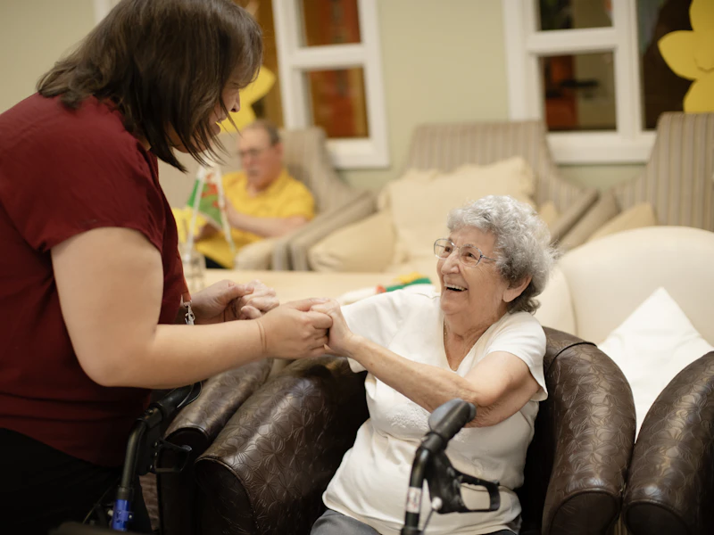 Une aide-soignante tient les mains d'une dame âgée assise dans son fauteuil.